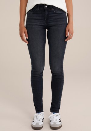 Dopasowane ciemnoniebieskie dżinsy noszone z białymi butami Adidas, na stojącej osobie na jednolitym tle.