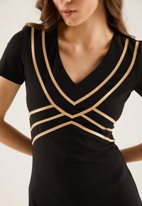 Svart klänning med V-ringning och korta ärmar, prydd med gyllene geometriska ränder över torso. Slät textur med en åtsittande design.