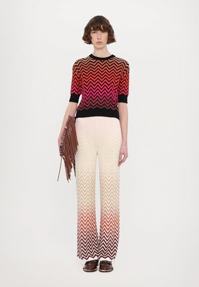 Pull en maille avec un motif en chevrons en rouge, rose et noir ; pantalon crème avec des détails en chevrons dégradés ; tenant une pochette frangée marron ; chaussures marron.