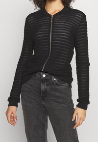 Woman wearing a black textured knit zip-up jacket with ribbed cuffs and faded black high-waisted jeans against a plain background.