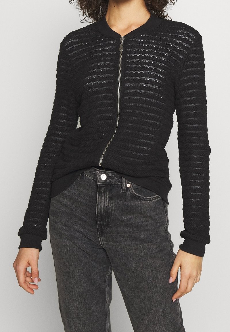 Woman wearing a black textured knit zip-up jacket with ribbed cuffs and faded black high-waisted jeans against a plain background.