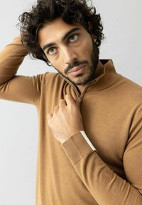 Hnedý pletený sveter s štvrťovým zipsom a rebrovanými manžetami s jemným bielym akcentom. Na ľavom rukáve je vyšívané logo. Jemná textúra.