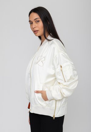 Frau mit langen dunklen Haaren, die eine weiße Satin-Bomberjacke mit goldenen Reißverschlüssen und einem bestickten "B" trägt, die Hände in den Taschen, nach links blickend.