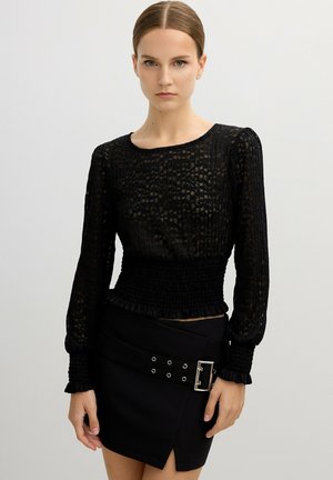 Top negro de mangas largas con patrón texturizado, detalles acanalados y cintura fruncida. Combinado con una falda negra que presenta herrajes de hebilla y acentos de ojal.