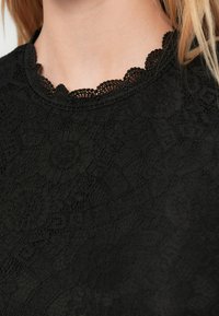 Top de renda preta com decote em formato de ondas. A textura intrincada apresenta padrões florais com um acabamento suave e elegante.