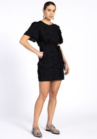 Schwarzes Kleid mit floraler Stickerei, kurzen Ärmeln und tailliertem Design. Getragen mit gemusterten Schwarz-Weiß-Schuhen. Einfaches Design, strukturierter Stoff.