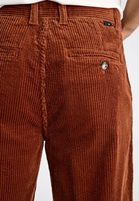 Pantalones de pana marrón con canalé vertical, con cierre de botón y bolsillos traseros. La textura parece suave y aterciopelada.