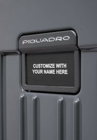 Étiquette de bagage grise affichant la marque "PIQUADRO" et une zone de texte personnalisable indiquant "PERSONNALISEZ AVEC VOTRE NOM ICI" en blanc. Fond texturé.