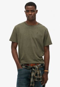 Camiseta de algodón verde oliva con cuello redondo, manga corta y un pequeño logo en el pecho. Combinada con jeans azules y una camisa de cuadros atada a la cintura.
