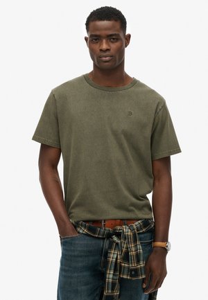 Olive green cotton t-shirt with a crew neckline, short sleeves, and a small logo on the chest. Paired with blue jeans and a plaid shirt tied at the waist.