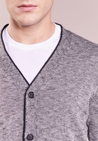 Närbild av en man som bär en ljusgrå stickad kofta över en vit T-shirt med rund halsringning, med två synliga mörka knappar på koftan.