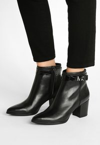 Schwarze Stiefeletten mit glänzender Oberfläche, spitzem Zehenbereich und blockiertem Absatz. Verfügt über einen glänzenden schwarzen Riemen mit Logo-Detail und einem seitlichen Reißverschluss.