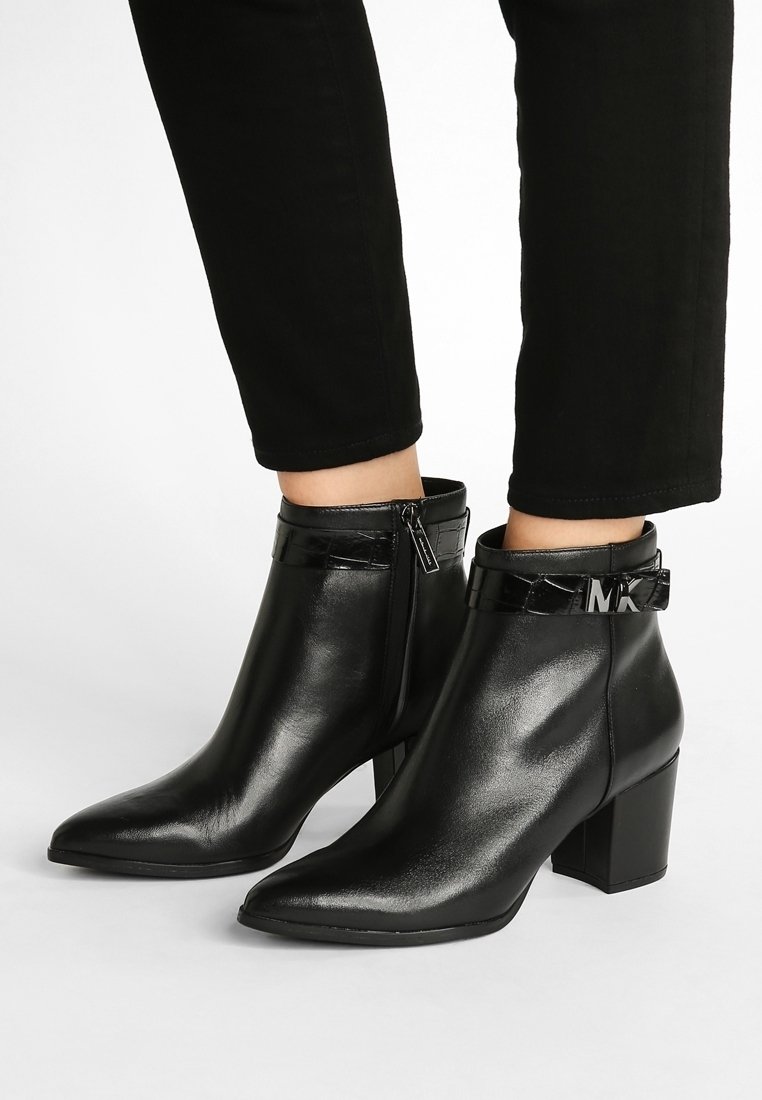 Schwarze Stiefeletten mit glänzender Oberfläche, spitzem Zehenbereich und blockiertem Absatz. Verfügt über einen glänzenden schwarzen Riemen mit Logo-Detail und einem seitlichen Reißverschluss.
