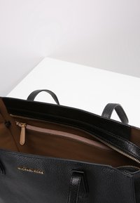 Sac fourre-tout en cuir noir grainé avec deux poignées supérieures, ferrures en or, poche intérieure zippée et doublure en tissu marron, avec un logo embossé à l'avant.