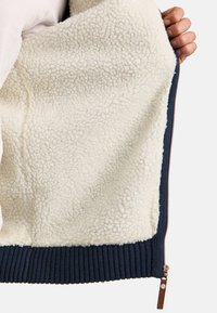 Fleece-lined jacket featuring a cream interior, ribbed navy cuff, and bronze zipper detail. Soft texture with a snug fit.