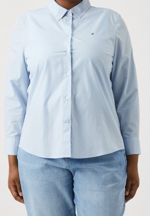 Chemise bleu clair à manches longues avec col et boutons, portée avec un jean bleu clair, montrant le torse et les bras sur un fond uni.