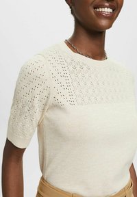 Kremowy sweter z krótkim rękawem, z teksturowanym, ażurowym wzorem na ramionach i gładkim korpusem. Posiada okrągły dekolt i ściągacze na mankietach.