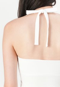 Bílý top na halter, s uvázáním na krku, s hladkou texturou a nízkým designem na zádech. Odkrytá pokožka je viditelná pod výstřihem.