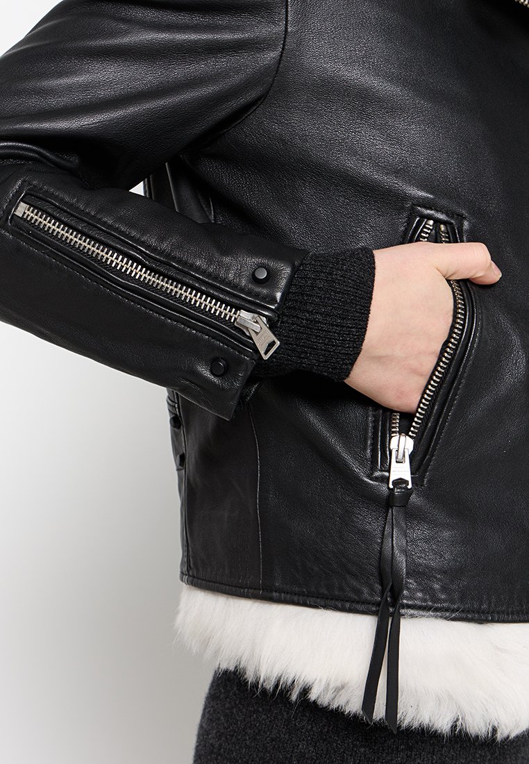 Schwarze Leder-Motorradjacke mit silbernen Reißverschlüssen, gerippten Strickbündchen und einem weißen Fellbesatz am Saum. Nahaufnahme des Taschenbereichs.