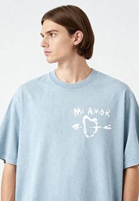 T-shirt oversize azzurro chiaro realizzato in morbido cotone, con una stampa grafica bianca di "Mi Amor" accompagnata da un design con cuore e freccia.