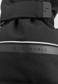 Giacca softshell nera con una texture morbida, caratterizzata da una linea di accento bianca orizzontale e la parola "SOFTSHELL" impressa su un nastro nero.