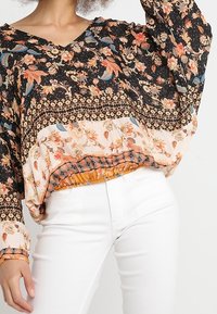 Femme portant un chemisier ample à motifs floraux noirs, beige et orange, associé à un pantalon blanc ajusté, sur un fond uni.