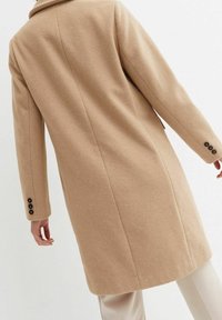 Manteau en mélange de laine beige avec une coupe droite, un col cranté et des boutons décoratifs sur les manches. Tissu texturé avec une finition lisse.