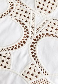 Tissu blanc présentant une broderie découpée complexe avec des motifs circulaires et floraux, des accents beiges et des détails de couture texturés.