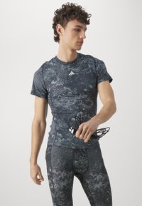 Svart og grå tettsittende treningstrøye med marmor mønster, har korte ermer og Adidas-logo på brystet. Modellen holder hodetelefoner.