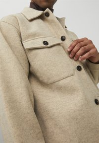 Beige wool blend jacket with a structured design, featuring two chest pockets, black buttons, and a ribbed brown collar. Soft texture.