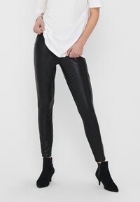 Black faux-leather leggings with a smooth, glossy finish, featuring a slim fit and ankle length. Paired with a white top and black ankle boots.