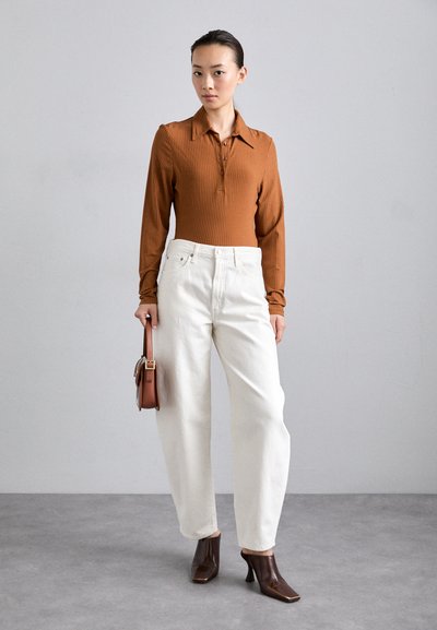 Haut marron à manches longues côtelé avec col, pantalon blanc à jambes larges et bottes à talons marron. Le mannequin tient un petit sac à main marron.