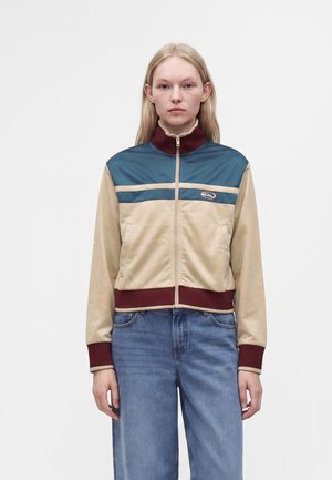Femme blonde debout portant une veste zippée à blocs de couleurs beige, bleu sarcelle et bordeaux avec un jean bleu sur un fond clair uni.
