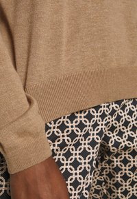 Bruine gebreide trui met geribbelde zoom, getoond naast een patroonbroek met een geometrisch ontwerp in roomkleurige en marineblauwe tinten.