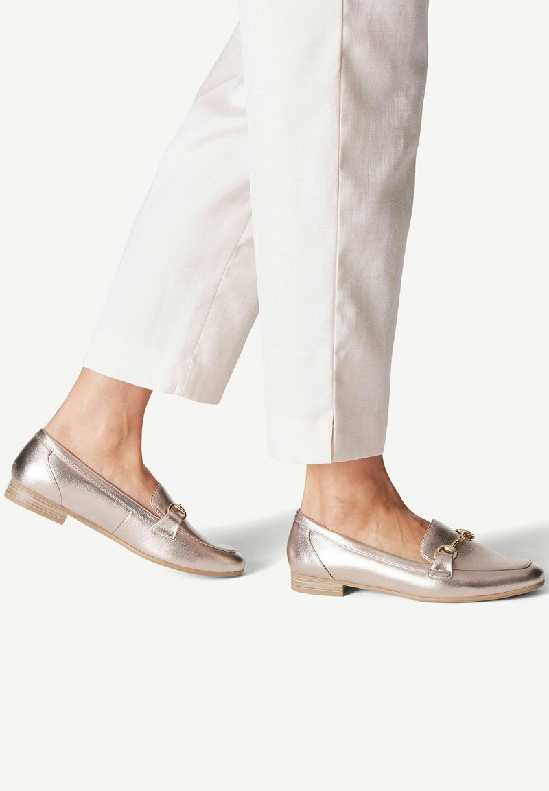 Kengät, joissa on pyöreä kärki, metallinhohtoiset kultaiset loaferit, joissa on koristeellinen solki ja matala korko, yhdistettynä vaaleisiin osittain leikattuihin housuihin.
