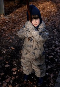 Bruine waterdichte overall met blauwe patronen, voorzien van een ritssluiting aan de voorkant, elastische manchetten en een capuchon. Het kind houdt een stok vast in een bosrijke omgeving.