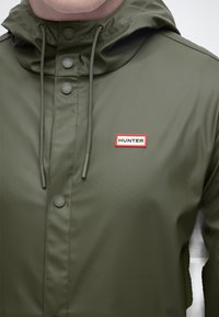 Αδιάβροχο μπουφάν σε ελιά πράσινο χρώμα με κουκούλα, κούμπωμα με κουμπιά, κορδόνια ρύθμισης και ορθογώνιο ένθετο λογότυπο "HUNTER" στο στήθος.