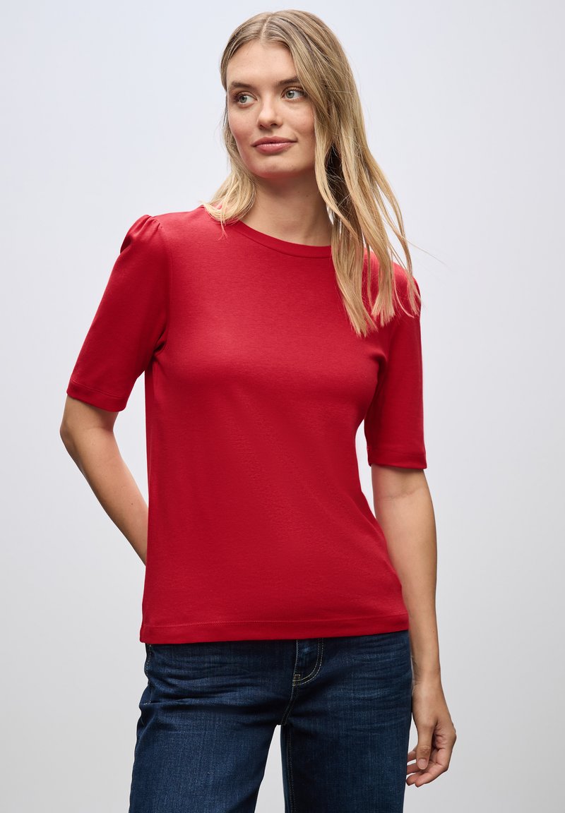 Rød kortærmet t-shirt med rund halsudskæring og let pufærmer, lavet af et glat stof. Båret sammen med blå jeans.