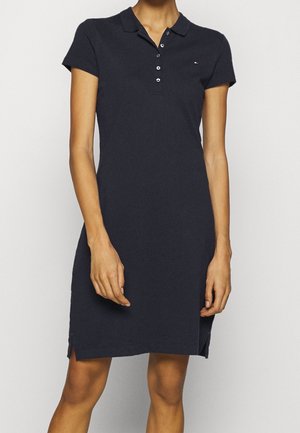 Femme portant une robe polo bleu marine foncé à manches courtes, longueur genoux, avec un col boutonné et un petit logo sur la poitrine.