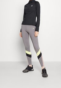 Černé dlouhé rukávy s zipem, v kombinaci se šedými legínami s geometrickými barevnými bloky v žluté, černé a růžové. Černé sportovní boty.