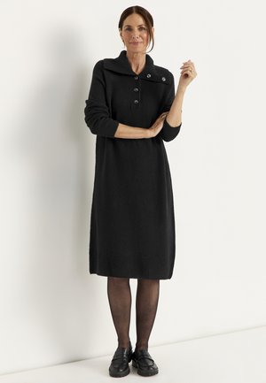 Woman standing against a wall wearing a black long-sleeve button-up sweater dress, sheer black tights, and black loafers, with hair tied back.