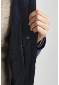 Navy blue jacket with a quilted interior, featuring a zippered pocket and snap closure. The fabric appears textured and lightweight.