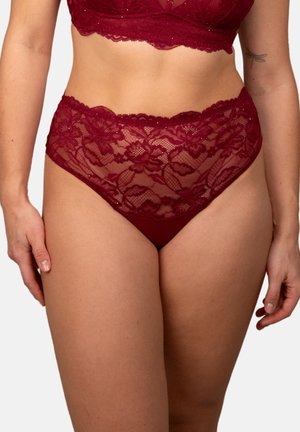 Woman wearing high-waisted red lace underwear and matching bra, showing midsection and upper thighs, with tattoo on left arm.