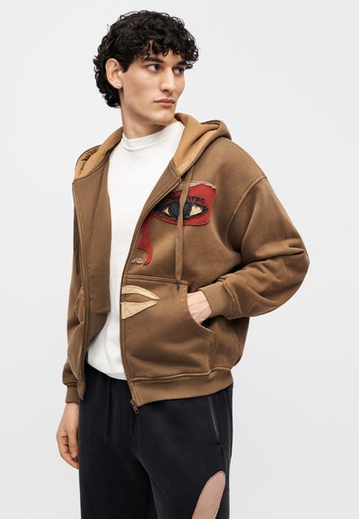 Καφέ φούτερ με φερμουάρ και σχέδιο ματιού και χειλιών σε κόκκινους και μπεζ αποχρώσεις. Μαλακό ύφασμα, τσέπες καγκουρό και κουκούλα με κορδόνι.