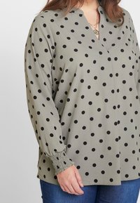 Blouse verte à pois noirs, décolleté en V, manches longues avec poignets smockés, tissu doux, coupe décontractée, associée à un jean bleu.
