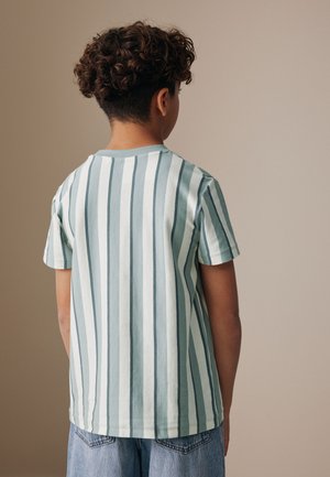 Göndör hajú fiú háttal áll, világoskék-fehér függőleges csíkos rövid ujjú inget és farmer sortot visel.