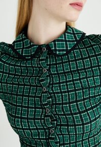 Groene jurk met een gestructureerd, gerimpeld ontwerp en een geblokte kraag. Het heeft een rij zwarte knopen aan de voorkant.