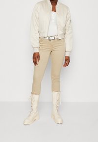 Osoba w kremowej skórzanej kurtce bomber, białym prążkowanym topie, beżowych obcisłych spodniach, białym pasku z srebrną klamrą oraz kremowych butach na sznurowadła.