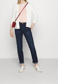 Cárdigan blanco, top a rayas naranja y blanco, vaqueros oscuros, zapatillas blancas con detalles rojos y un pequeño bolso bandolera rojo.