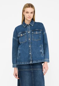 Giacca di jeans blu con chiusure a bottone, due tasche sul petto e colletto classico. Vestibilità leggermente ampia e dettagli cuciti.